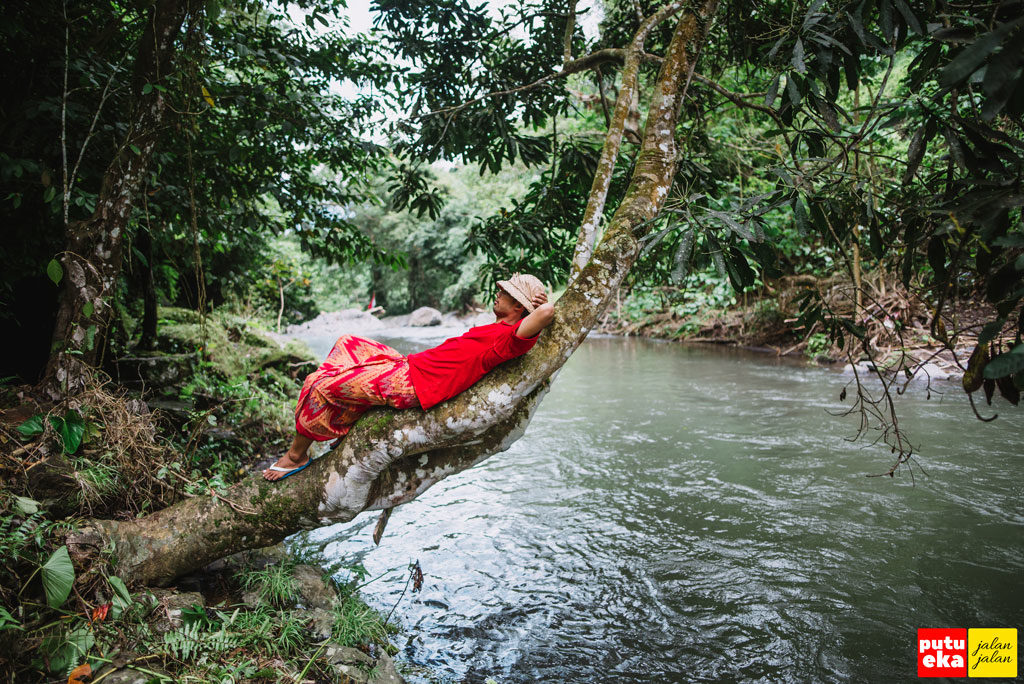Putu Eka Jalan Jalan sedang beristirahat di batang pohon yang menjorok ke tengah sungai